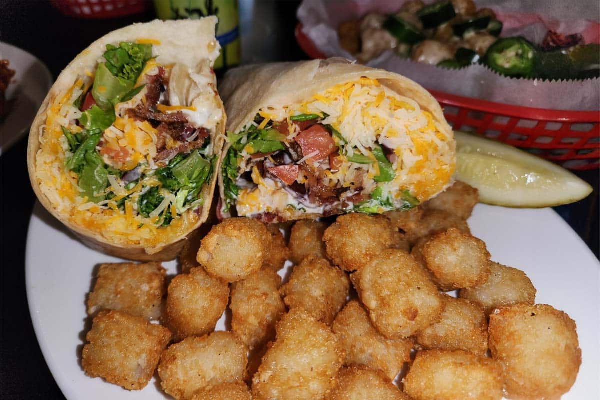 Brewski’s Club Wrap Platter with Tater Tots A large club wrap with bacon and cheese from Brewski's, cut in half and served on a white plate with a side of crispy tater tots and a pickle spear.