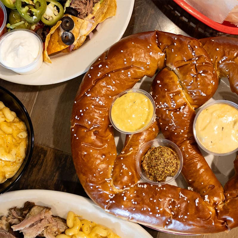 A food spread at Brewski's featuring a giant soft pretzel with cheese and mustard, loaded nachos, and mac and cheese on a wooden table.