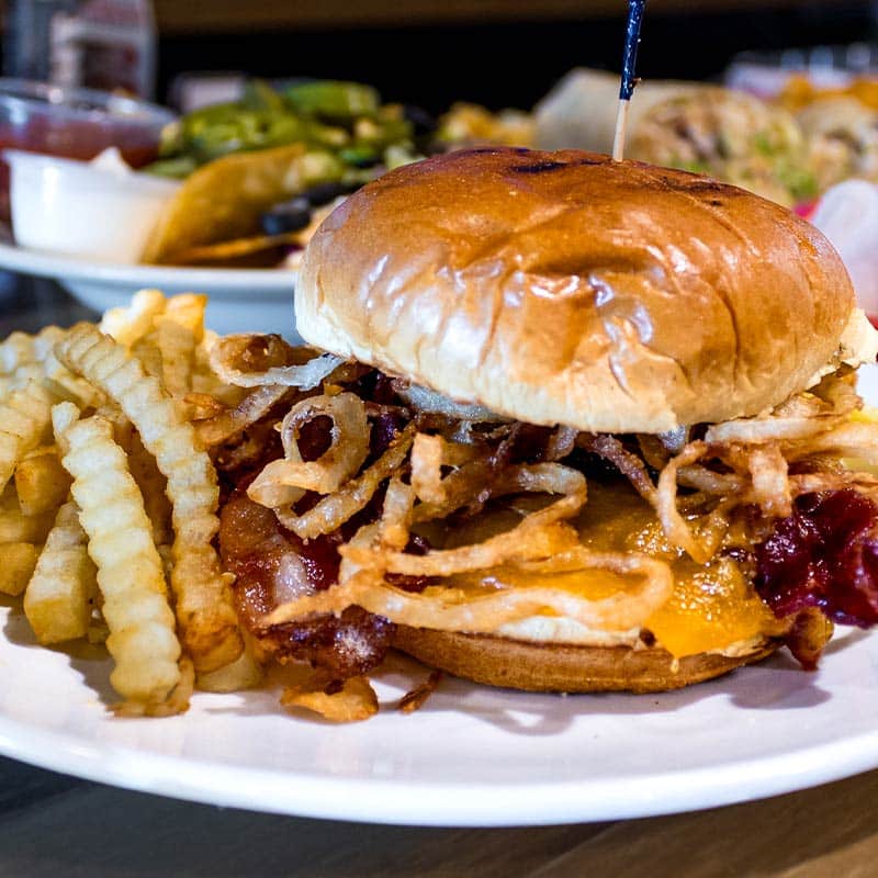 A close-up of a Brewski's BBQ bacon cheeseburger topped with crispy fried onions, served on a white plate with a side of golden crinkle-cut fries.