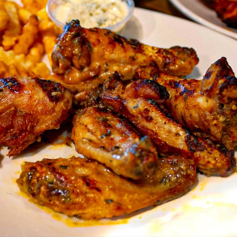 A close-up of Brewski's signature grilled chicken wings, seasoned and saucy, served on a white plate with crinkle-cut fries and a side of creamy dipping sauce.