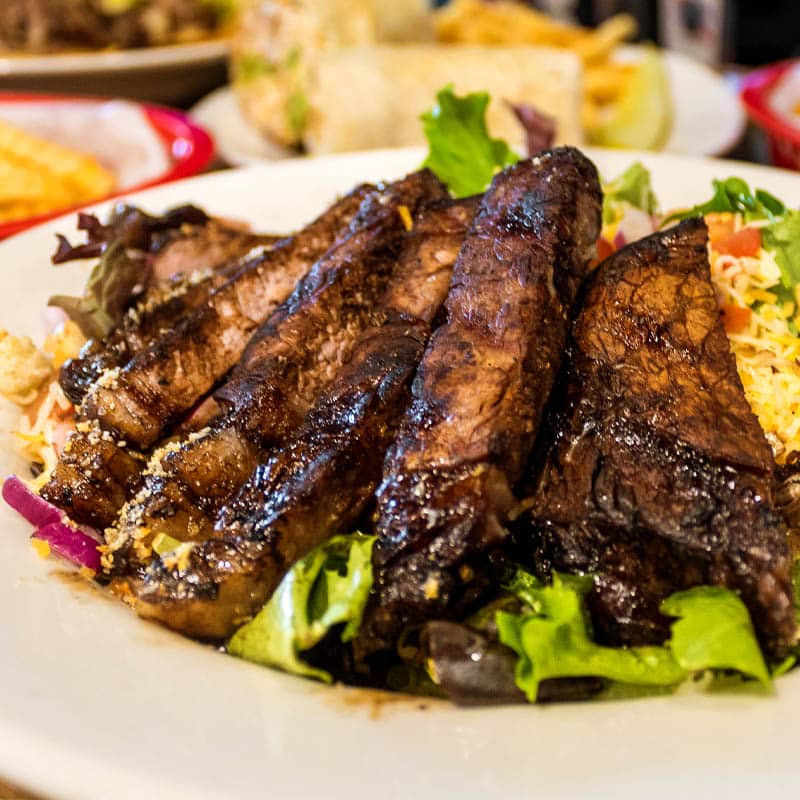 A grilled steak salad from Brewski's, with thick-cut charred steak slices served over a bed of mixed greens, red onions, and tomatoes on a white plate.